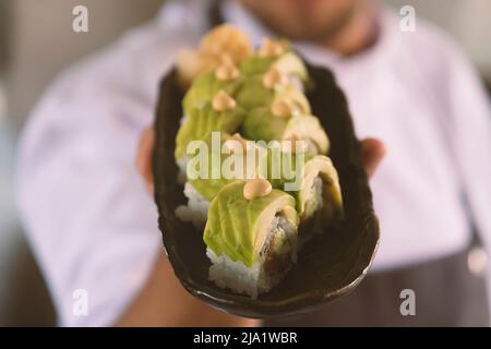 Der junge Koch hält einen Teller Sushi mit Avocado und Lachs. Stockfoto
