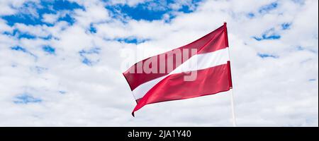 Die lettische Flagge winkt im Wind. Flagge Lettlands auf wolkig weißem Himmel Hintergrund. Stockfoto