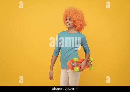 Fröhliches egoistisches Kindermädchen in lockiger Perücke hält Penny Board, Hipster Stockfoto