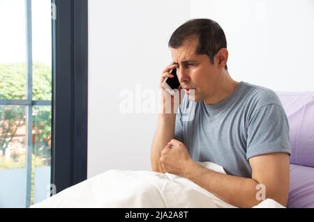 Foto eines kranken Geschäftsmannes, der auf dem Bett sitzt und den Arzt im Schlafzimmer anruft Stockfoto