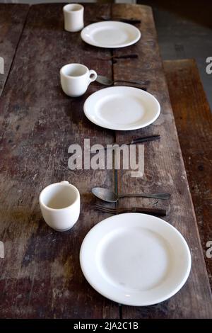 Auf einem abgenutzten rustikalen Holztisch ruht antikes Tischgestellen mit schlichten weißen Tassen und Tellern und angeschlassenem Besteck. Stockfoto