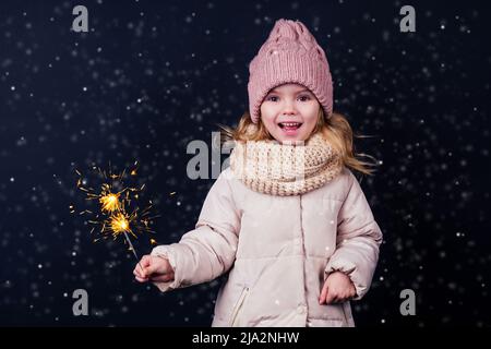 Charmantes kleines Mädchen in einem gestrickten rosa Hut hält Feuerwerk auf schwarzem Hintergrund in einem Studio.Cute blonde Kind mit Weihnachten Dream.Happy Kind genießen die Stockfoto