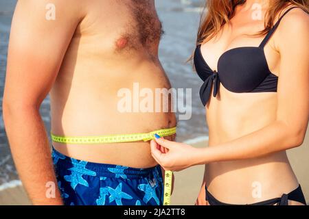 Schlanke Frau misst die Volumina eines fetten Mannes am Strand Stockfoto