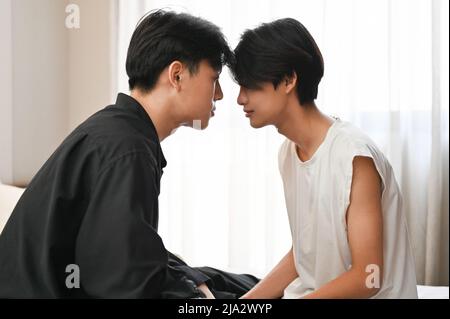 Zwei hübsche junge schwule asiatische Männer entspannen sich, sitzen von Angesicht zu Angesicht auf dem Bett und zeigen eine gewisse Zuneigung zu LGBT. Schwule Freunde Konzept Stockfoto