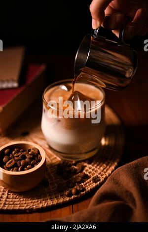 Ein Barista gießt einen kalten Brühkaffee oder dunklen Kaffee in ein Glas Eis mit frischer Milch. Kaffeebild über dunklem Hintergrund. Erfrischungsgetränke Stockfoto