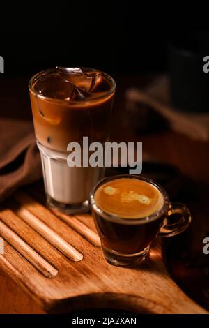 Ein Glas vereisten Latte und ein Schuss heißen Espresso auf einem rustikalen Holzbrett auf dunklem Hintergrund. Nahaufnahme, Café-Getränkekarte. Stockfoto