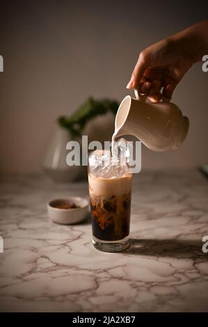 Nahaufnahme, die Hand der Barista gießt eine frische Milch in ein Glas Eiskaffee und macht einen Eislatte. Stockfoto