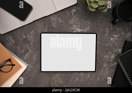 Ein digitales Tablet-Touchpad mit weißem Bildschirm und Bürozubehör auf grunge Zementstruktur auf grauem Hintergrund. Modernes Arbeitsplatzkonzept. Draufsicht, Stockfoto