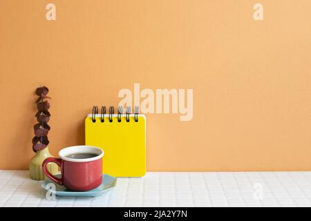 Tasse Kaffee und Notizblock, trockener Eukalyptus auf weißem Tisch. Orangefarbener Wandhintergrund. Platz zum Kopieren Stockfoto