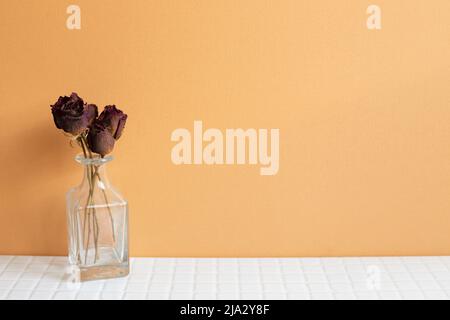Vase mit roten trockenen Rosenblüten auf weißem Tisch. Orangefarbener Wandhintergrund. Kopierraum Stockfoto