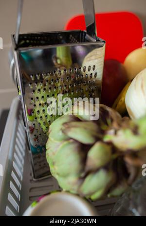 Gereinigtes Gemüse und Geschirr auf grauem Kunststoffschüssel-Trockengestell Closeup Artichoke Reflexion in silberner Reibe auf dem Geschirrtrocknungsgestell mit anderem Gemüse Stockfoto