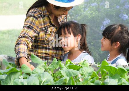 Junge Mutter im Strohhut, die ihre Töchter im Garten unterrichtete. Das kleine Mädchen hilft ihrer Mutter im Garten, einer kleinen Gärtnerin. Nettes Mädchen Pflanzen Stockfoto