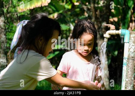 Zwei kleine Schwestern waschen sich die Hände am Wasserhahn im Freien. Zwei niedliche Mädchen drehen den Wasserhahn zu, um sich im Park die Hände zu waschen. Stockfoto