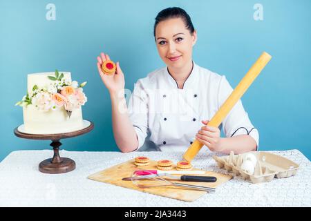 Konditorei Konditorin Bäckerin Frau kochte cremig weißen zweistufigen Hochzeitstorte (Geburtstag) mit frischen Blumen auf dem Tisch und Cookies im Studio auf blau Stockfoto