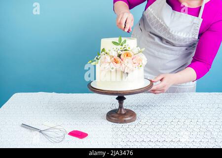 Die Konditorin (Konditorin) schmückt appetitlich cremig-weißen zweistufigen Hochzeitstorte, der mit frischen Blumen auf einem Tisch mit Spitze verziert ist Stockfoto