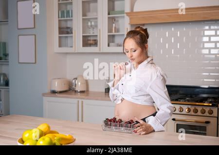 Gesunde Ernährung Schwangerschaft und Menschen Konzept Schwangere junge Frau, die reife Kirschen in der Küche zu Hause trägt weißes Hemd und lächelt glücklich. Kaukasische werdende Mutter genießt diesen Zustand. Stockfoto