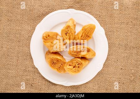 Mehrere Scheiben süßer Honig-Baklava mit einer weißen Keramikschale auf Sackleinen, Nahaufnahme, Draufsicht. Stockfoto