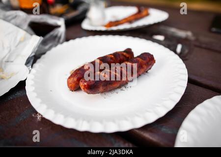 Nahaufnahme von zwei gegrillten Würstchen auf weißem Pappteller auf braunem Holztisch mit Tellern, Plastiktüte, im Hintergrund ein Stück Papier Stockfoto