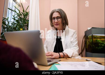 Angenehme ältere kaukasische Frau mit Laptop, arbeitet im Modedesign-atelier, erstellen Kleidung Skizzen auf Holztisch gelegt. Kreativer Beruf, onl Stockfoto