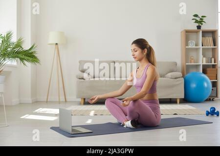 Eine athletische junge Frau meditiert vor dem Laptop und hört ruhige Musik oder Affirmationen. Stockfoto