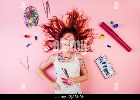 Fun schmutzige Künstler Maler Flecken Flecken Flecken von Farbe auf dem Gesicht liegend auf dem Boden neben Palette, Tube Farbe, Pinsel auf einem rosa Hintergrund Stockfoto