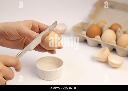 Eine weibliche Hand hält ein Hühnereier und bricht es mit einem Messer. Papptablett mit Eiern. Konzentrieren Sie sich auf das Ei. Stockfoto