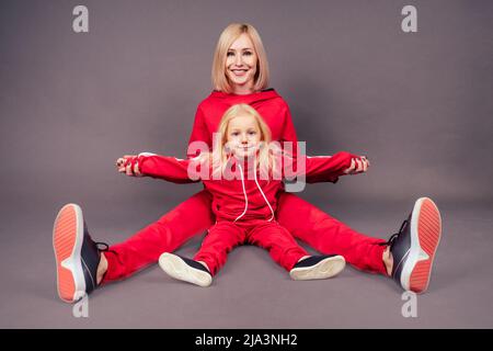 In voller Länge Mutter mit Tochter im Sport suits.beautiful junge blonde Frau mit einem kleinen Mädchen sitzt auf dem Boden in rot stilvollen Anzug Studio schwarz Stockfoto