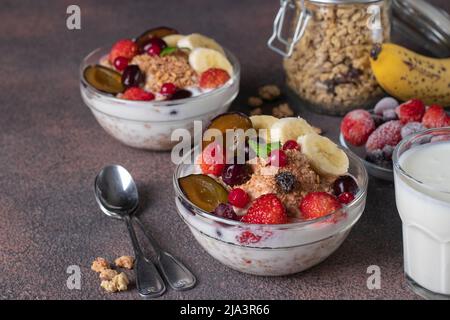 Müsli knusprig mit Naturjoghurt, gefrorenen Beeren und Früchten und Nüssen in zwei Glasschüsseln auf braunem Hintergrund Stockfoto