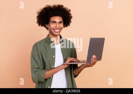 Porträt von attraktiven fröhlich qualifizierte wellig-haarige Kerl Agent Broker mit Laptop Entwicklung isoliert über beige Pastellfarben Hintergrund Stockfoto