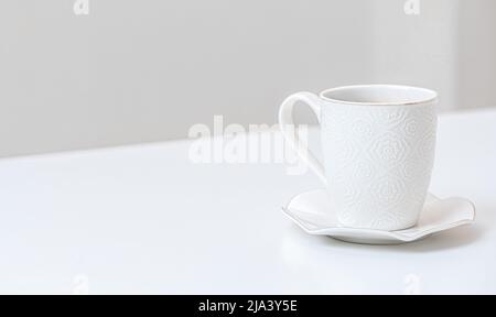 Weißer Porzellanbecher auf einem Tisch im Innenbereich. Weiße Untertasse und Tasse Kaffee auf weißem Tisch. Keramik-Teetasse auf weißem Küchentisch. Seitenansicht Stockfoto