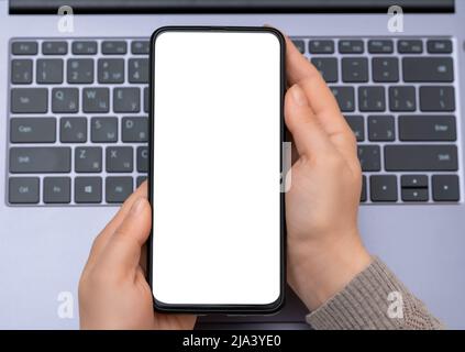 Hände von Frau mit Handy mit leerem Bildschirm auf Laptop-Tastatur Hintergrund. Draufsicht Mockup-Bild der Hände halten weißen Bildschirm Handy Stockfoto