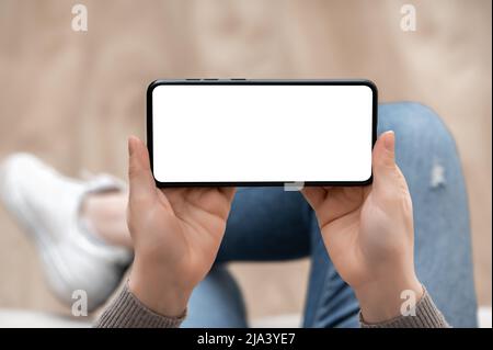 Mockup-Bild von oben, in dem die Frau das Mobiltelefon horizontal mit einem weißen Bildschirm hält. Frau in Jeans sitzt mit gekreuzten Beinen und hält das Handy horizontal Stockfoto