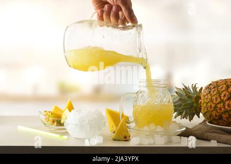 Person, die gekühlten Ananassaft aus dem Krug auf der Küchenbank mit Obst und Eis serviert. Vorderansicht. Horizontale Zusammensetzung. Stockfoto