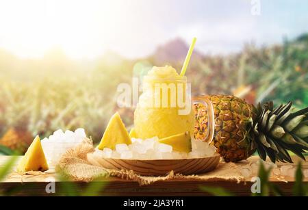 Ananas-Slush mit Früchten um ihn herum auf einem Holztisch draußen mit Ananaspflanzen auf dem Feld. Vorderansicht. Horizontale Zusammensetzung. Stockfoto