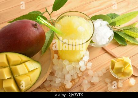 Detail eines Glases mit Mangosaft mit Eis auf einem Holztisch mit Früchten und Würfeln drum herum. Draufsicht. Horizontale Zusammensetzung. Stockfoto