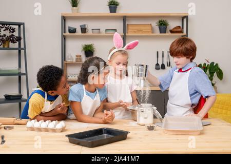 Kinder gießen Teig zusammen, tragen Schürzen und schauen sich mit einem breiten Lächeln an, während sie appetitliche Kekse für ihre Eltern zubereiten Stockfoto