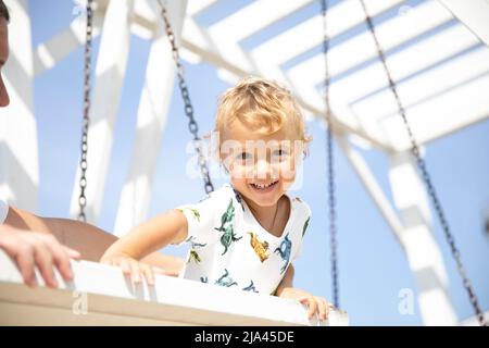 Kleiner Junge, 3 Jahre alt, blonde, am sommerlich warmen sonnigen Tag in einem T-Shirt, spaziert im Park und reitet mit einem fürsorglichen Erwachsenen auf einer Schaukel unter einer weißen Pergala. Stockfoto