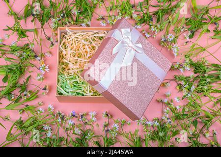 Rosa Geschenkbox mit Schleife auf rosa Hintergrund in einem Rahmen aus Wildblumen. Stockfoto