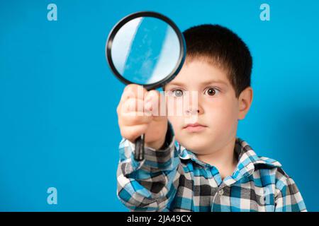 Ein Vorschuljunge hält in der Hand eine Lupe auf einem blauen Hintergrund mit einem Platz für Text. Stockfoto