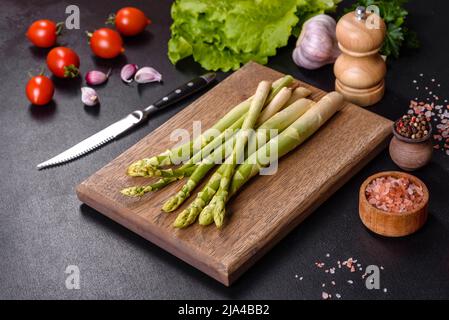 Ein Haufen Zweige von frischem grünem rohem Spargel auf einem hölzernen Schneidebrett vor einem dunklen Betongrund Stockfoto