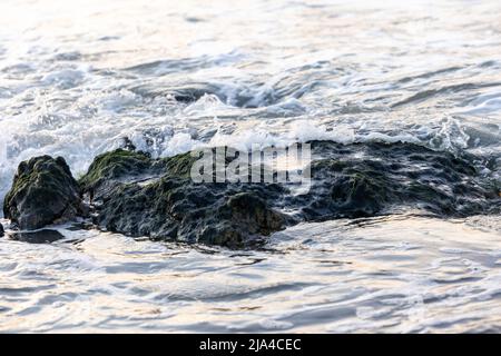 Brechende Wellen zu den großen Felsen am Ufer, die etwas weißen Meeresschaum und kleine Blasen erzeugen, mit ein wenig Reflexion des Sonnenuntergangs im Wasser. Cap Blanc-Nez, Frankreich. Hochwertige Fotos Stockfoto