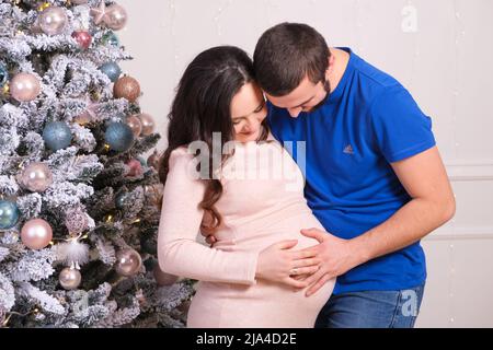 Ein glücklicher Ehemann und eine glückliche Frau bereiten sich darauf vor, Eltern zu werden und Spaß am Weihnachtsbaum zu haben. Schwangerschaft und Weihnachten Stockfoto