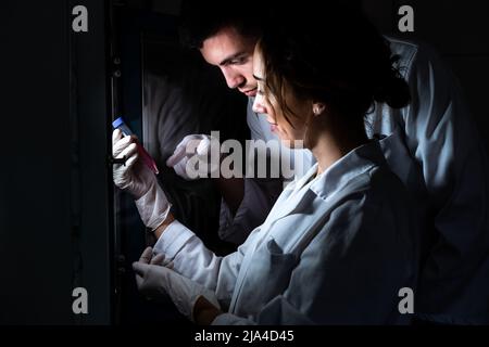 Zwei lächelnde Wissenschaftler im Labormantel, die einen mittleren Schlauch mit Zellen halten und studieren. Forschung, Impfstoff, Gesundheit Stockfoto