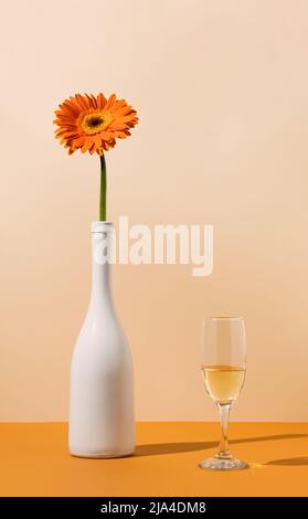 Weiße Champagner-Flasche mit oranger Gänseblümchen-Blume und Glas auf dem Tisch. Minimale Federzusammensetzung. Retro-Style. Stockfoto