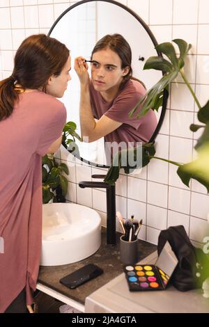 Nicht-binäre Trans-Frau tut ihre Augenbraue Make-up Blick in den Spiegel im Badezimmer zu Hause Stockfoto