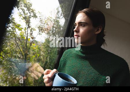 Nicht-binäre Transfrau mit Kaffeetasse und Blick aus dem Fenster zu Hause Stockfoto