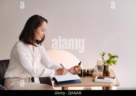 Das Aromatherapeutin-Mädchen sitzt in ihrem Büro und hält eine Flasche aromatisches Öl in den Händen und schreibt etwas auf. Es gibt ätherisches Öl Stockfoto