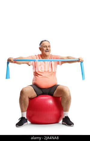Ein glücklicher, reifer Mann in Sportkleidung sitzt auf einem Fitnessball und trainiert mit einem auf weißem Hintergrund isolierten Widerstandsband Stockfoto
