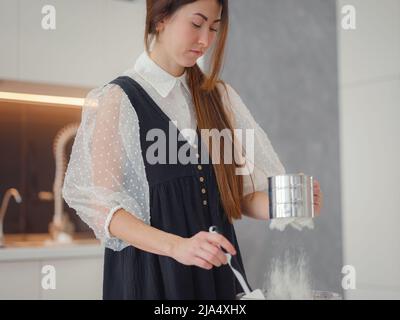 DIY Hausmannskost Konzept. Brunette Frau sieben Mehl für Kuchen durch Sieb, salzen Teig. Junge Frau lächelt, liebt es, in schönen modernen k kochen Stockfoto
