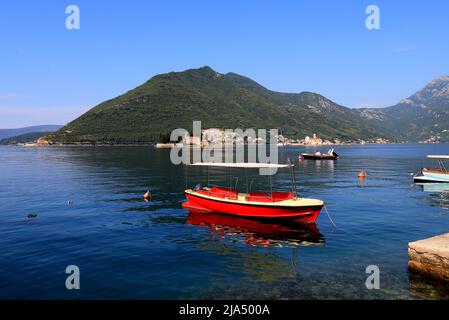 Boka Kotor, Montenegro. Ein rotes Boot fährt entlang der Bucht Kotor vor dem Hintergrund der berühmten Insel und der Kirche Madonna am Riff in der Nähe der Stadt Perast Stockfoto